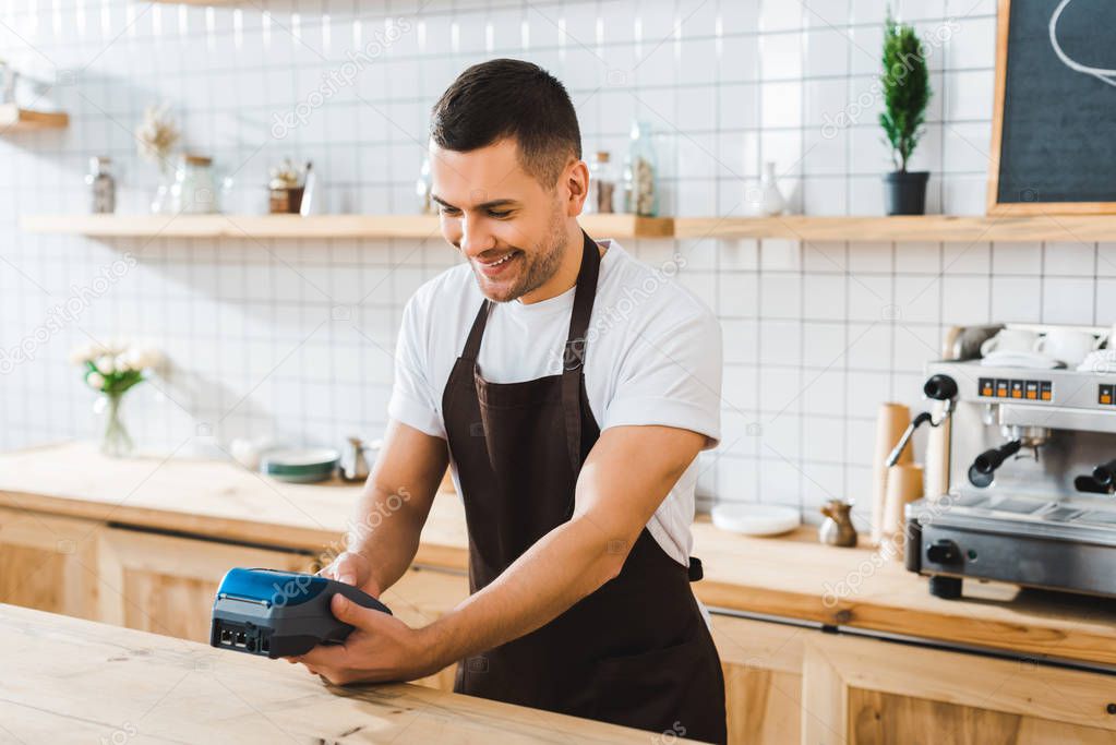 handsome cashier standing near bar counter in brown apron and holding blue terminal in coffee house