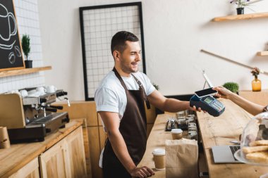 kahverengi önlük sayacında bar yakınındaki yakışıklı barista ayakta ve terminal wile kadın smartphone coffee House ile ödemek tutarak