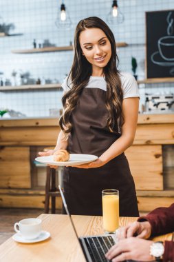 selective focus of attractive waitress holding plate with caucasian near man sitting at table with laptop and smiling in coffee house clipart