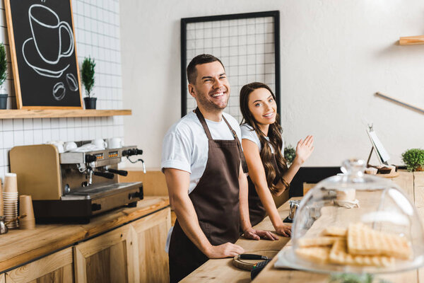 cashiers in aprons standing and smiling behind bar counter in coffee house