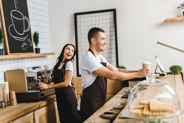 handsome cashier giving paper cups wile attractive barista making coffee and smiling behind bar counter in coffee house