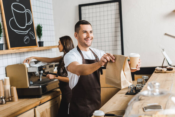 handsome cashier giving paper cup and bag wile barista making coffee behind bar counter in coffee house