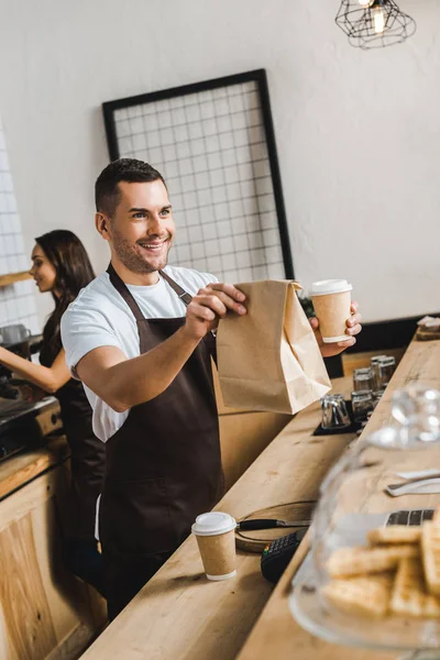Handsome Cashier Brown Apron Giving Paper Cup Bag Wile Barista — Stock Photo, Image