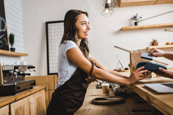 attractive cashier standing in brown apron and holding terminal wile man paying with credit card in coffee house