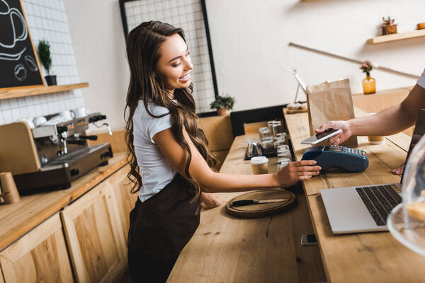 attractive cashier standing in apron and holding terminal wile man paying with smartphone in coffee house