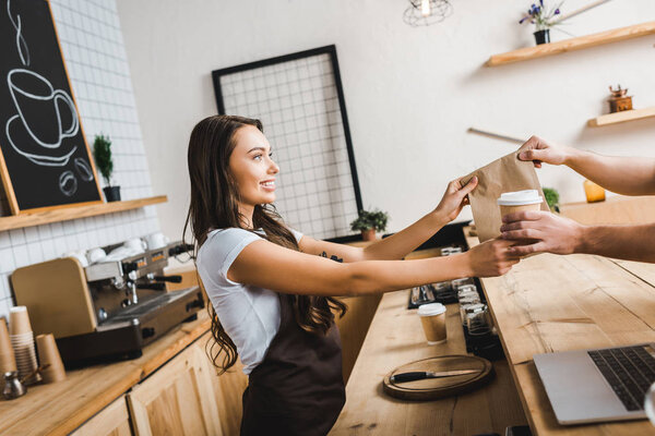 attractive cashier in brown apron giving paper cup and bag to man in coffee house