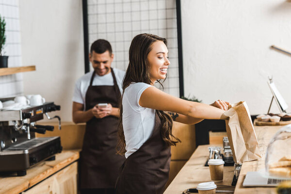attractive cashier in brown apron packing paper bag wile barista looking to smartphone near bar counter in coffee house
