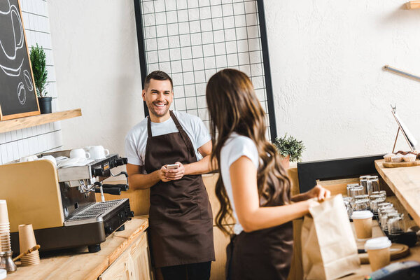 selective focus of handsome cashier with smartphone and barista standing behind bar counter in coffee house
