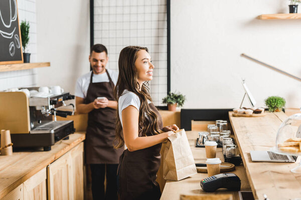 selective focus of attractive cashier with paper bag and barista with smartphone standing behind bar counter in coffee house