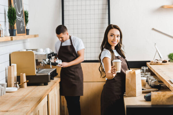 attractive smiling cashier with paper cup with barista in brown apron making coffee behind bar counter in coffee house
