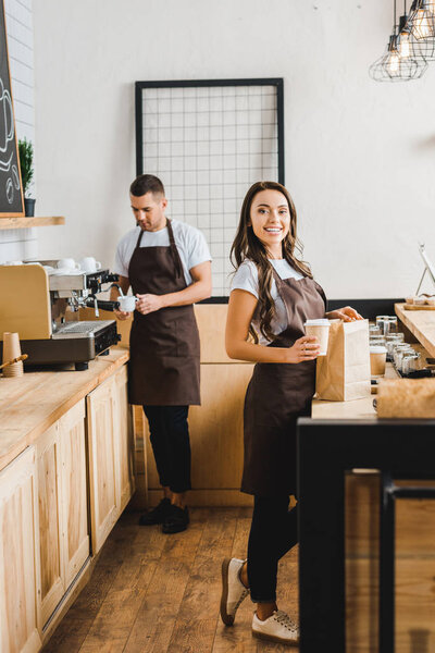 attractive cashier with paper cup and bag with barista in brown apron making coffee behind bar counter in coffee house