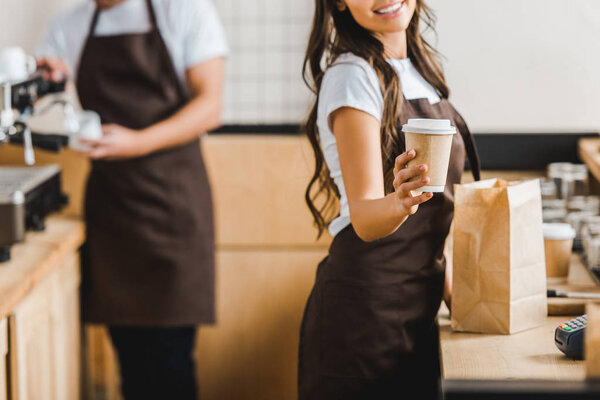 cropped view of cashier with paper cup and bag with barista in brown apron in coffee house