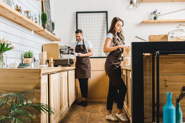 attractive cashier with paper bag and barista making coffee behind bar counter in coffee house