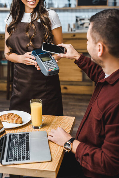 waitress holding terminal wile freelancer at table paying with smartphone in coffee house