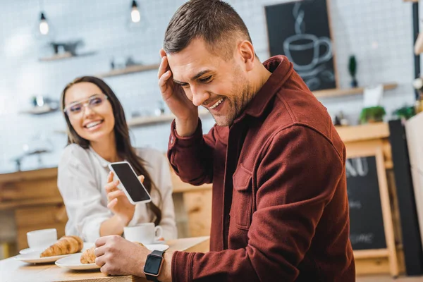 穿着勃艮第衬衫的帅哥坐在桌旁 笑着迷人的黑发女人 在咖啡屋展示智能手机的空白屏幕的选择性焦点 — 图库照片