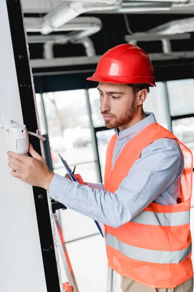 Pensive Fireman Helmet Checking Fire Alarm While Holding Clipboard — Stock Photo, Image