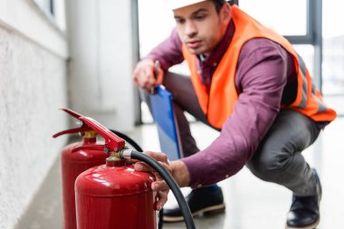 selective focus of red extinguishers near fireman in helmet holding clipboard