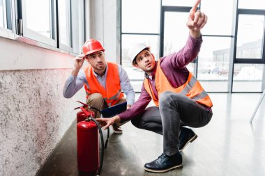 handsome fireman pointing with finger near coworker while sitting near red extinguishers  clipart