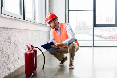 serious fireman in helmet holding clipboard while looking at extinguishers  clipart