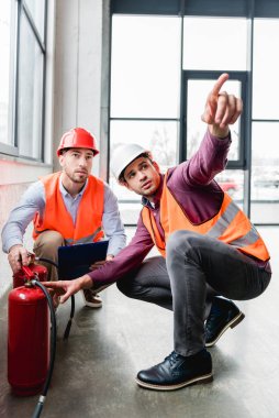 fireman pointing with finger near coworker while sitting near red extinguishers  clipart