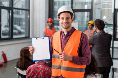 selective focus of cheerful fireman pointing at blank clipboard with pen near coworkers giving talk on briefing on background clipart
