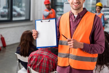 cropped view of cheerful fireman pointing at blank clipboard with pen near coworkers giving talk on briefing on background clipart