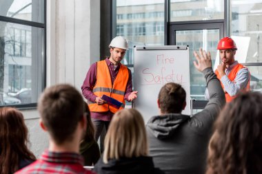 selective focus of handsome firemen in helmets giving talk on briefing near white board with fire safety lettering and looking at man raising hand clipart
