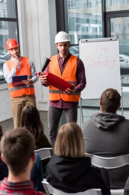 selective focus of handsome firefighters in helmets standing near white board with fire safety lettering and holding extinguisher and clipboard in hands clipart