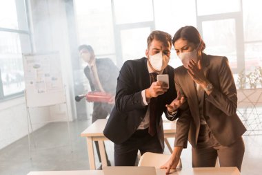 seçici odak içinde yazıhane duman ile smartphone kullanarak maskeli dehşete meslektaşlarının