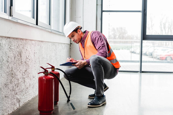 handsome fireman in helmet sitting near extinguishers and holding clipboard