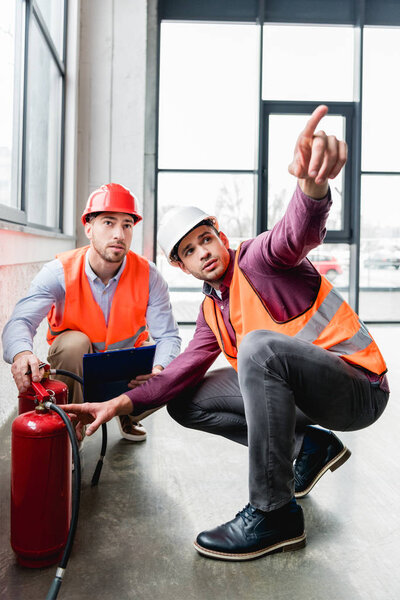 fireman pointing with finger near coworker while sitting near red extinguishers 