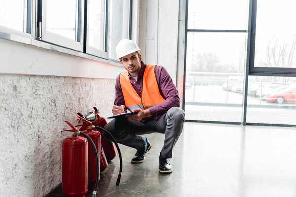 serious fireman in helmet holding clipboard and pen while looking at camera near extinguishers 