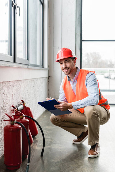 cheerful fireman in helmet holding clipboard and pen while looking at camera near extinguishers 