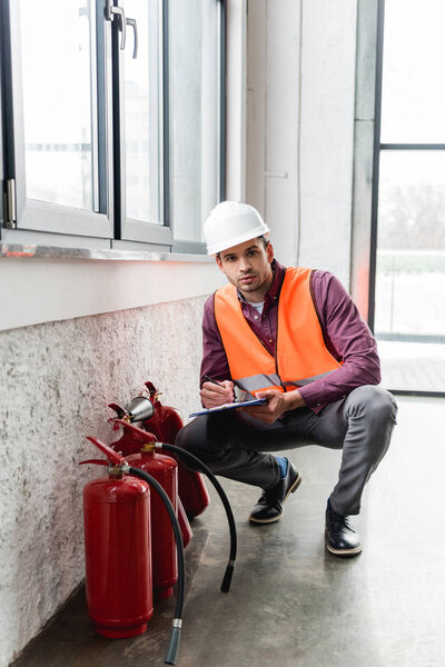 handsome fireman in helmet holding clipboard and pen while looking at camera near extinguishers 