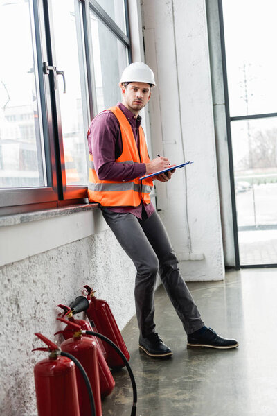 handsome fireman in helmet and uniform holding clipboard and pen while standing near extinguishers 