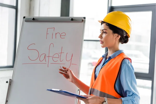 female firefighter in helmet holding clipboard and pen while talking near white board with fire safety lettering