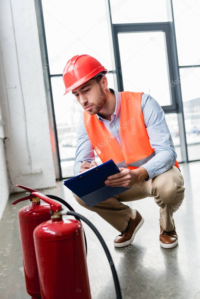 handsome fireman in helmet checking extinguishers while sitting with clipboard