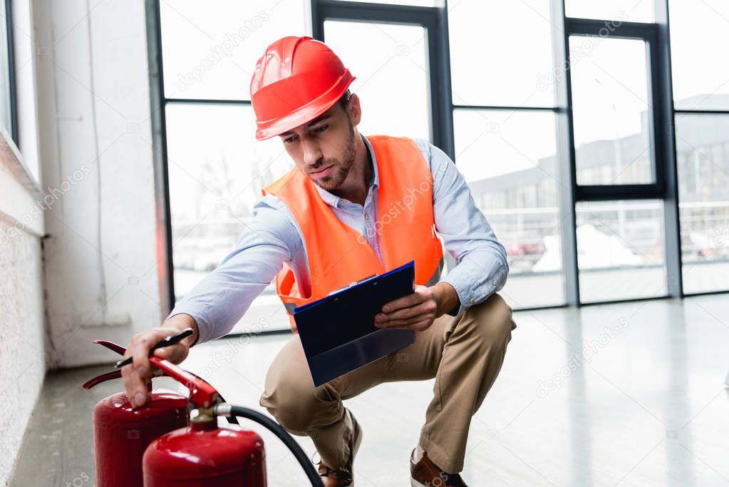 serious fireman in helmet checking extinguishers while sitting with clipboard