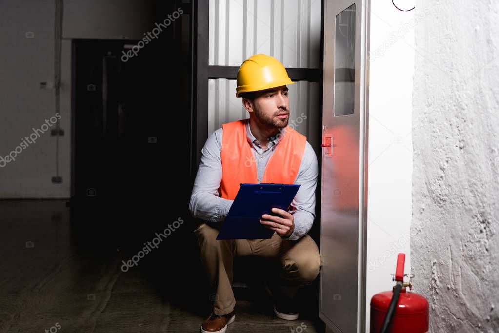 fireman in helmet looking at fire panel while sitting and holding clipboard