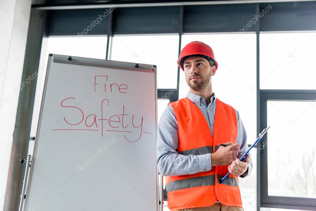 handsome firefighter in helmet holding clipboard and pen while standing near white board with fire safety lettering
