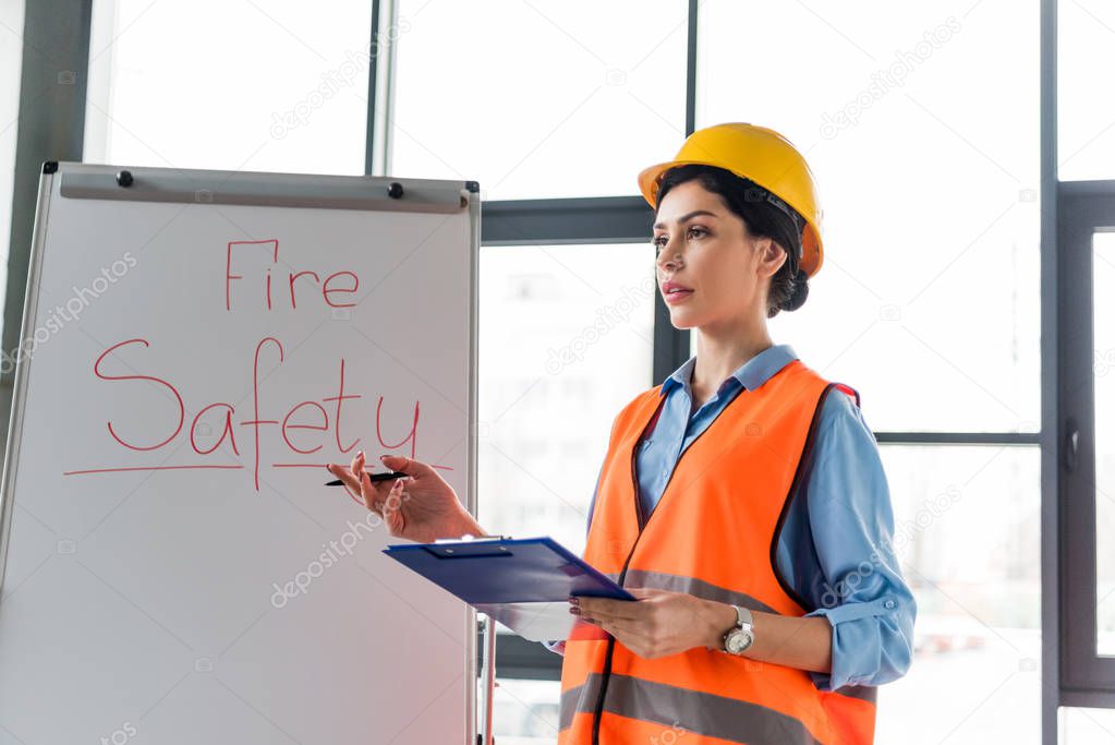 female firefighter in helmet holding clipboard and pen while standing near white board with fire safety lettering