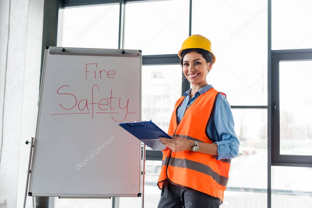 happy female firefighter in helmet holding clipboard and pen while standing near white board with fire safety lettering