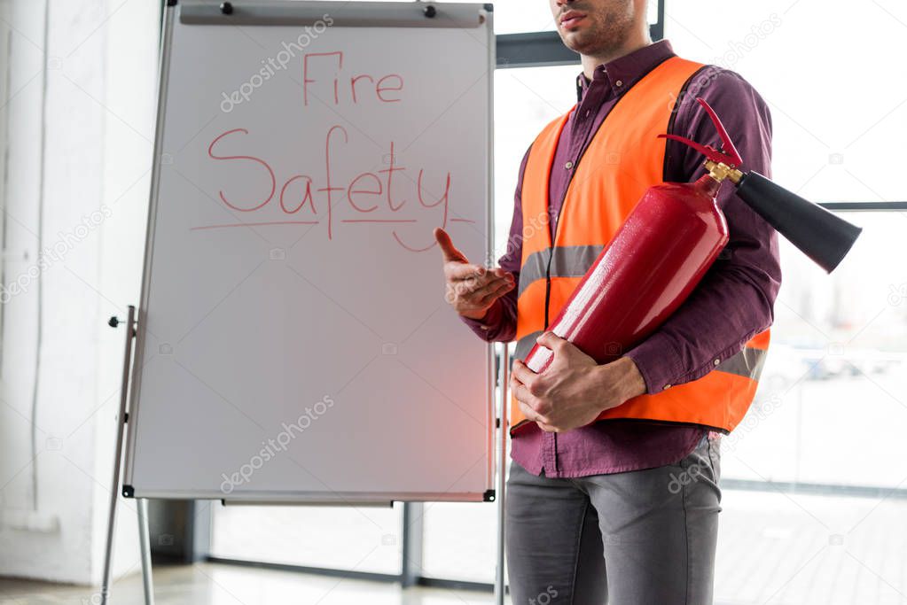 cropped view of fireman holding red extinguisher while standing near white board with fire safety lettering