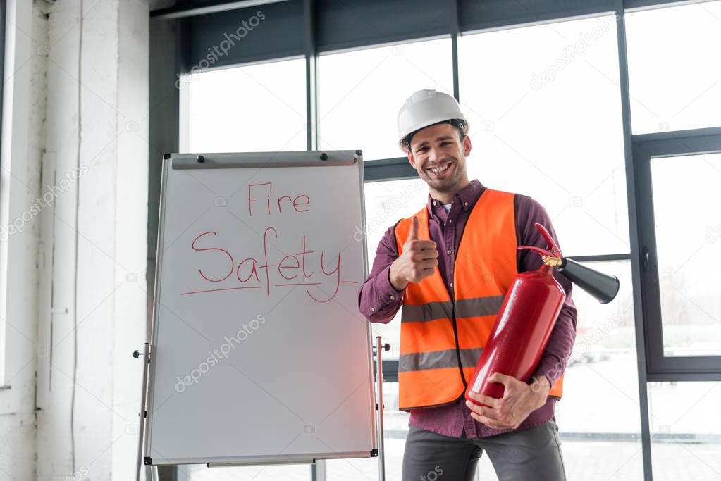 cheerful fireman holding red extinguisher and showing thumb up while standing near white board with fire safety lettering