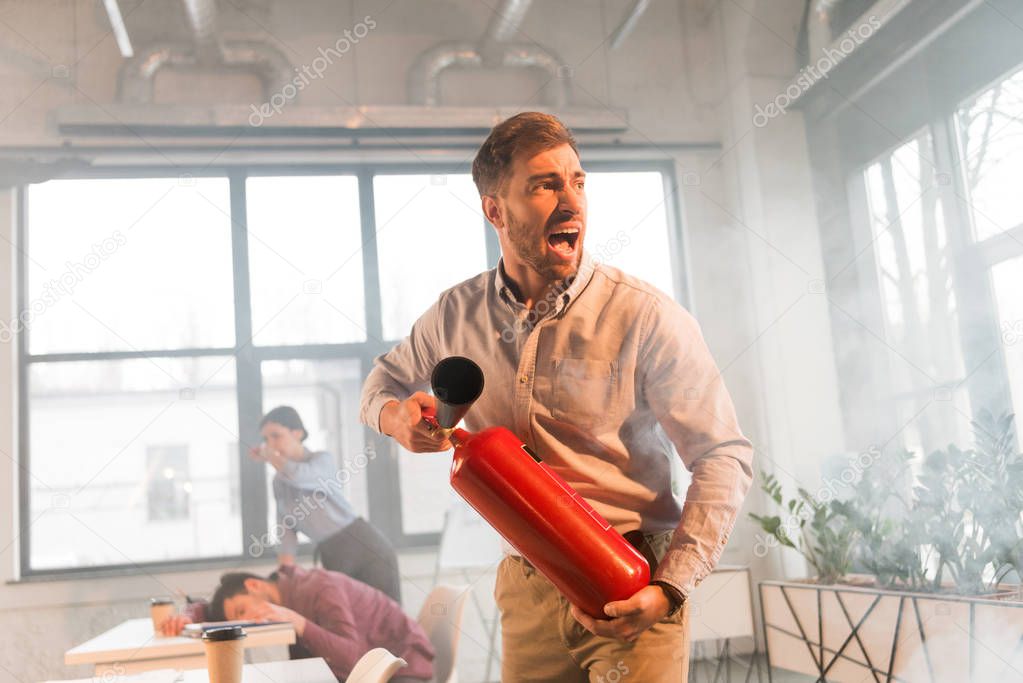 frightened businessman holding extinguisher and screaming in office with smoke near coworkers