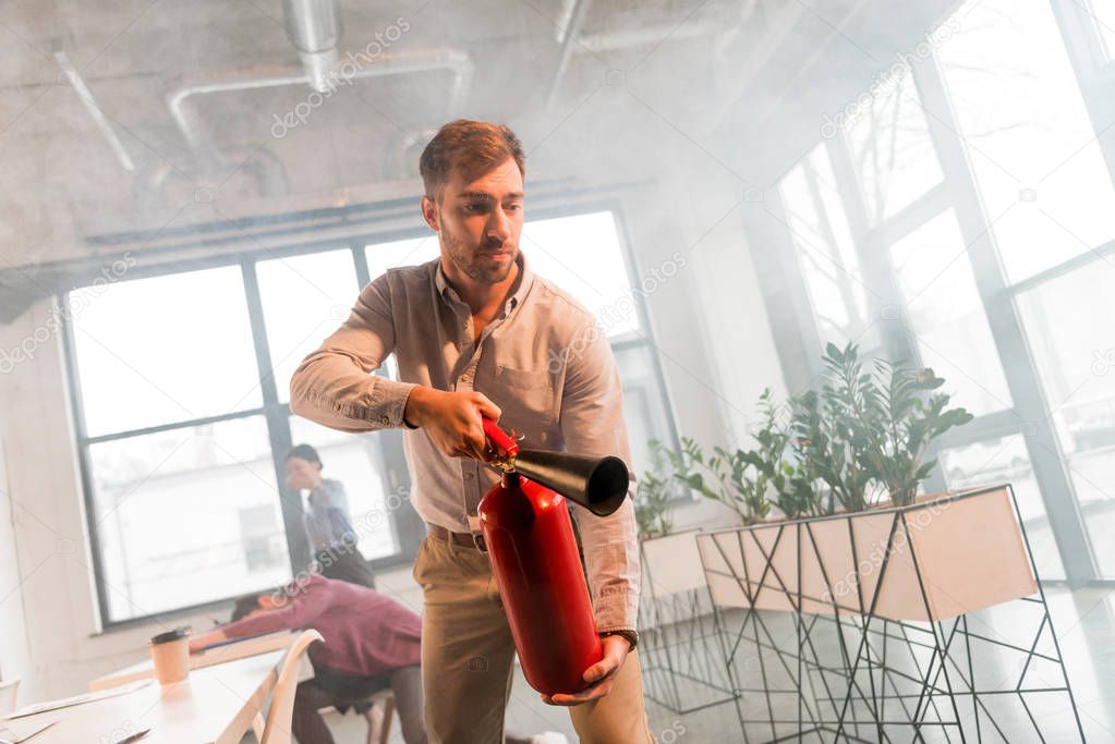 handsome businessman holding extinguisher in office with smoke near colleagues