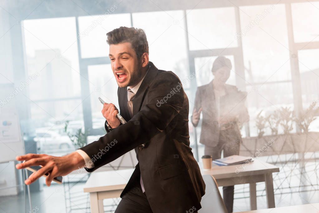 terrified businessman holding smartphone and screaming in office with smoke near female coworker