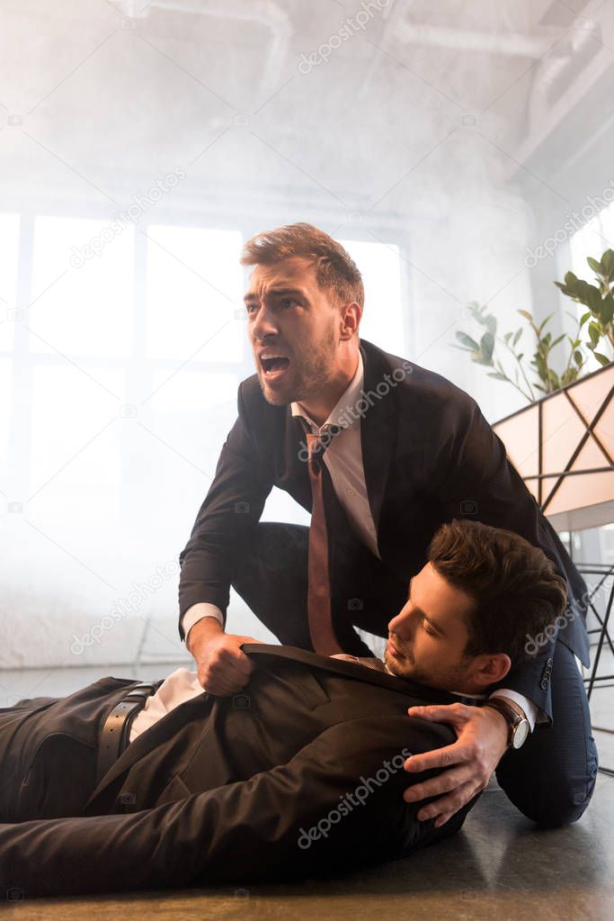 scared businessman screaming near colleague lying on floor in office with smoke