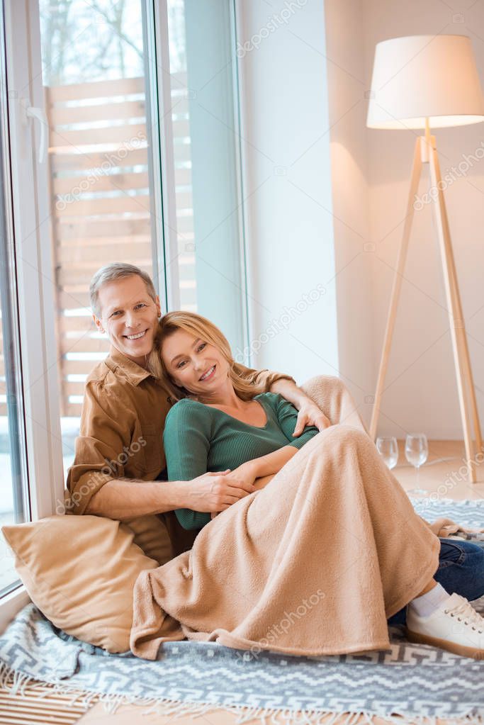 happy husband hugging wife sitting together on floor by large window 