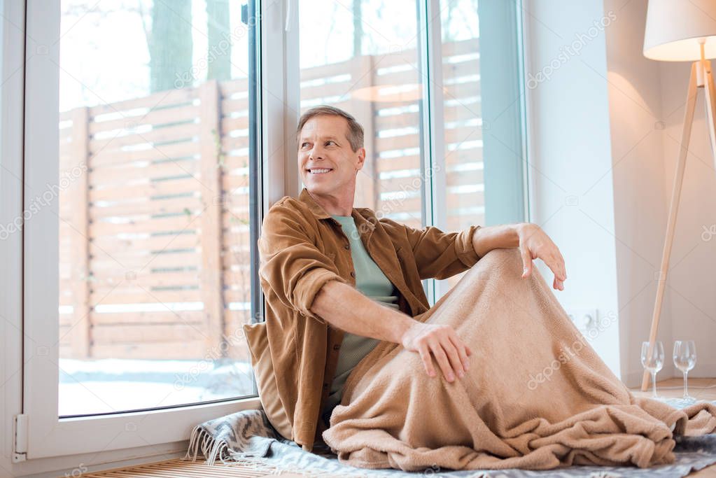 happy man sitting on floor under soft fleece blanket and looking through large window